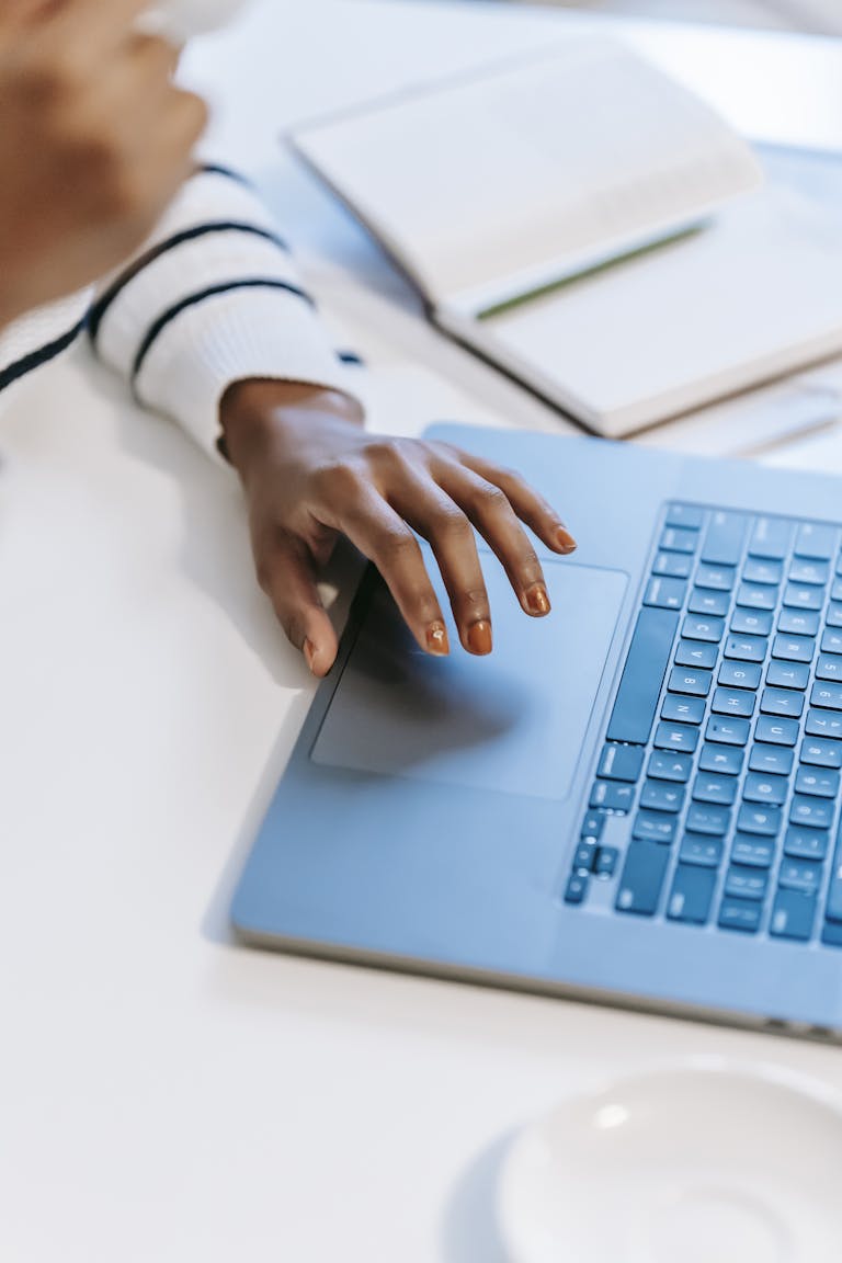 High angle of crop anonymous ethnic woman working remotely at white table on laptop near notebook with pen while drinking coffee in light workplace